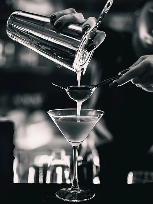 Czarno-białe, artystyczne zdjęcie przedstawiające dłonie barmana, który precyzyjnie przelewa koktajl z shakera przez sitko barmańskie do eleganckiego kieliszka typu martini.