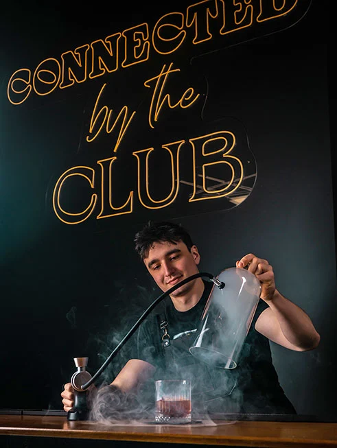 Uśmiechnięty barman w czarnym fartuchu przygotowujący efektowny wędzony koktajl pod szklanym kloszem na tle świecącego neonu "Connected by the Club" w Klub58.