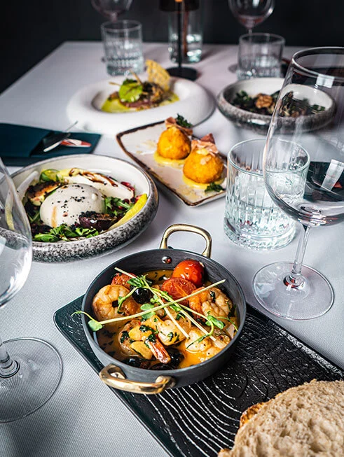 Zastawiony stół w restauracji Klub58 z gorącymi krewetkami w żeliwnym naczyniu na pierwszym planie, sałatką z burratą, smażonymi krokietami i kieliszkami do wina.
