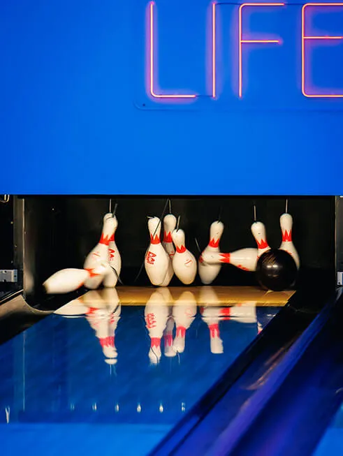 Dynamiczny moment uderzenia czarnej kuli do bowlingu w biało-czerwone kręgle, które rozpraszają się na końcu lśniącego toru. Nad maszyną widoczny jest fragment świecącego neonu z napisem "LIFE", całość w niebieskim oświetleniu Klub58.