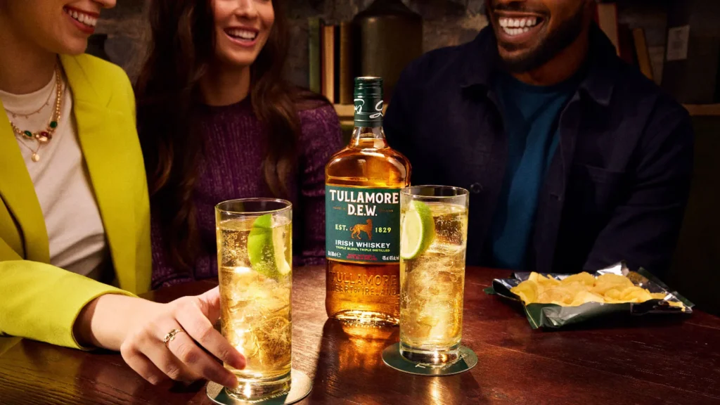 Grupa uśmiechniętych znajomych siedzących przy drewnianym stole, na którym stoi butelka irlandzkiej whiskey Tullamore D.E.W., dwa orzeźwiające drinki z lodem i limonką oraz paczka chipsów.