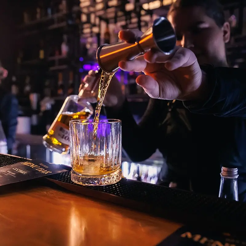 Barman w Klub58 precyzyjnie nalewający z metalowego miarki (jiggera) bursztynowy alkohol do kryształowej szklanki z lodem. Szklanka stoi na czarnej macie barowej obok menu, na drewnianym blacie w nastrojowo oświetlonym lokalu.