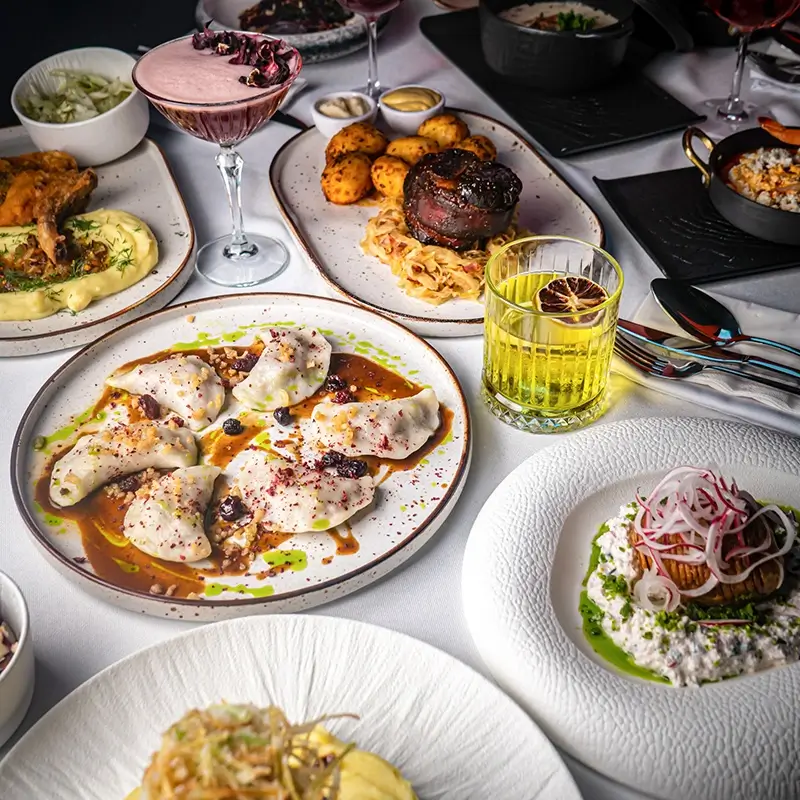 Bogato zastawiony stół w restauracji na białym obrusie, na którym widać m.in. talerz apetycznych pierogów w ciemnym sosie, porcję pieczonego mięsa z ziemniakami, eleganckie nowoczesne przystawki oraz kolorowe koktajle barowe.