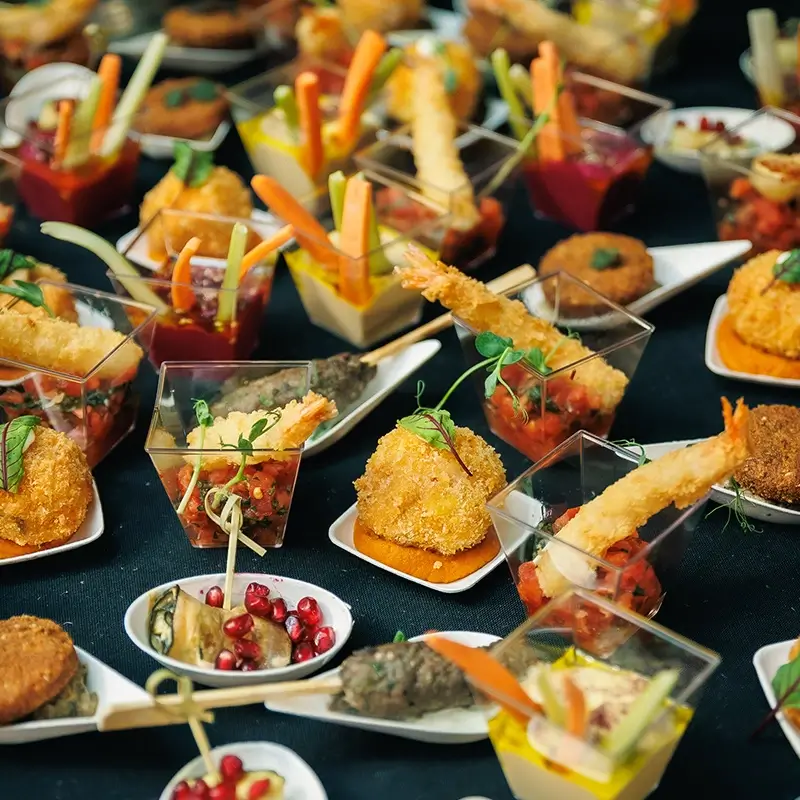 Bogaty bufet z eleganckimi przekąskami typu finger food na ciemnym tle, obejmujący m.in. chrupiące krewetki w tempurze w małych pucharkach, złociste krokiety, słupki świeżych warzyw z dipem oraz roladki posypane pestkami granatu.
