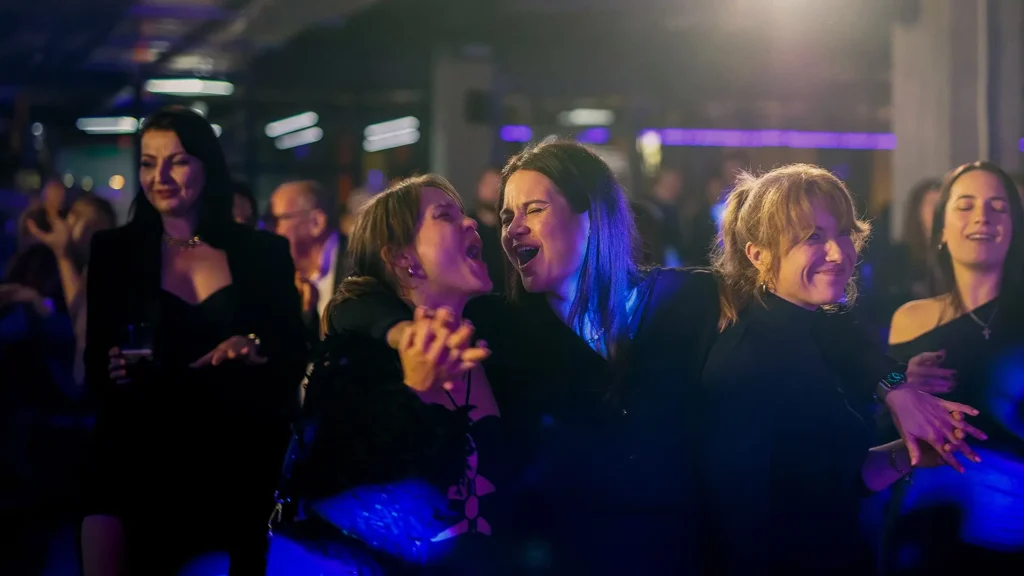 Grupa radosnych, śpiewających kobiet bawiących się wspólnie w nastrojowo oświetlonym, klubowym wnętrzu. Zdjęcie promujące wydarzenie z okazji Dnia Kobiet w Klub58.