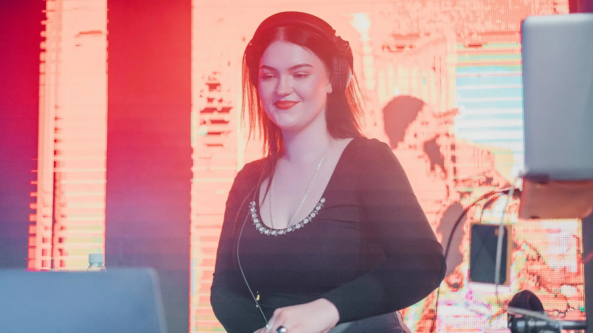 Uśmiechnięta kobieta, DJ WI.ZA, ze słuchawkami na głowie, miksująca muzykę za konsoletą didżejską. Artystka ubrana jest w czarną bluzkę z ozdobnym dekoltem, a w tle widać jasno oświetlony ekran LED w czerwono-różowych, klubowych barwach.