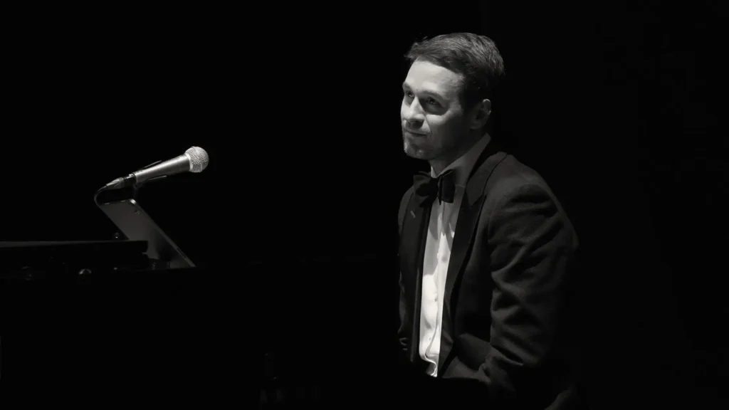 Czarno-białe, nastrojowe zdjęcie przedstawiające pianistę i wokalistę Jacka Szwaja. Artysta jest ubrany w elegancki smoking z muszką i siedzi przy instrumencie klawiszowym, a przed nim znajduje się statyw z mikrofonem.