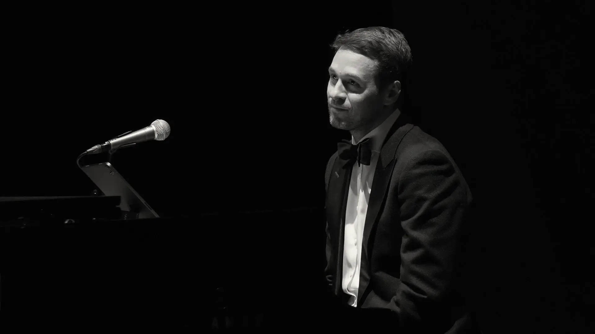 Czarno-białe, nastrojowe zdjęcie przedstawiające pianistę i wokalistę Jacka Szwaja. Artysta jest ubrany w elegancki smoking z muszką i siedzi przy instrumencie klawiszowym, a przed nim znajduje się statyw z mikrofonem.