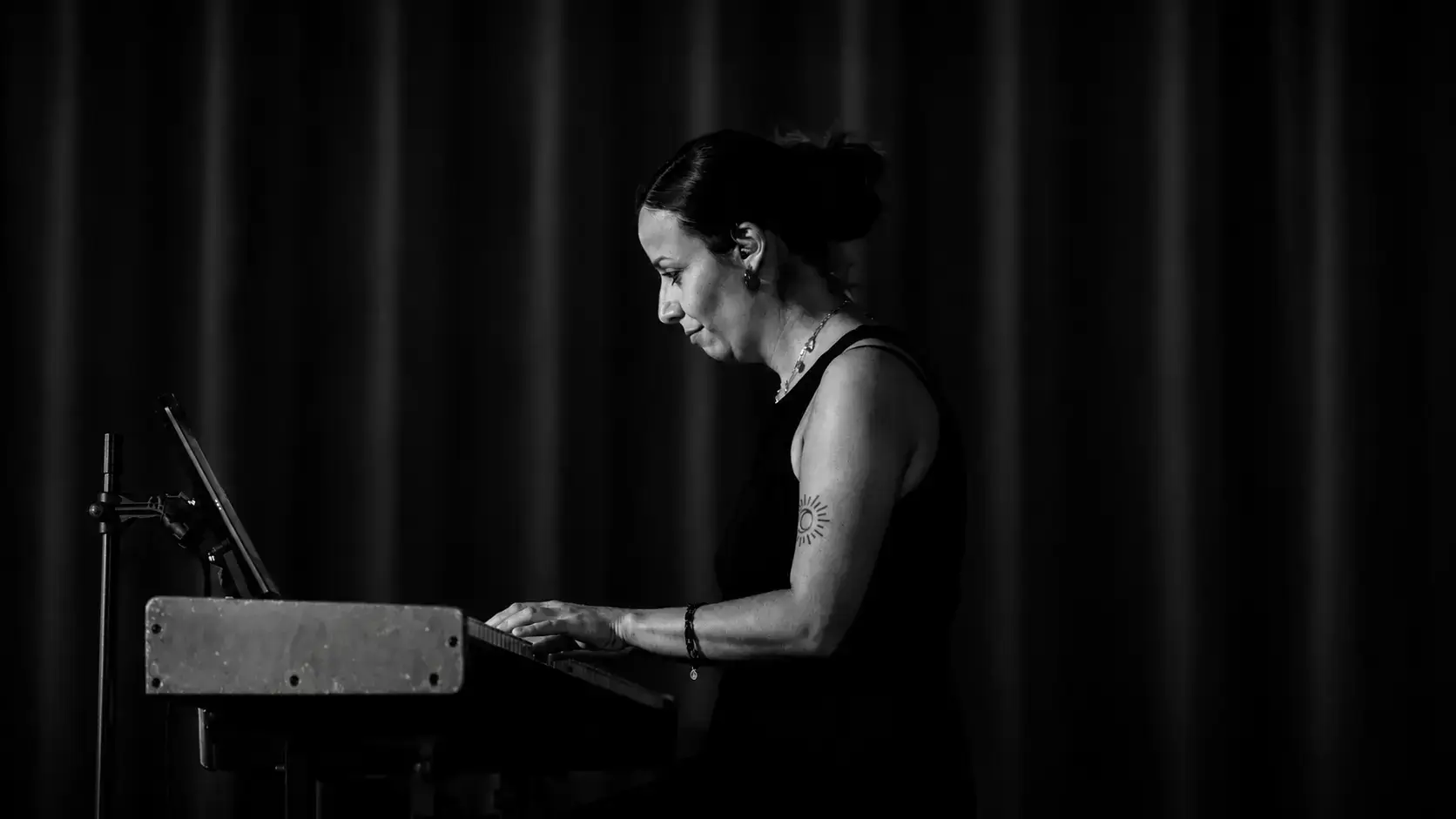 Czarno-biały plakat promujący koncert zespołu Maja Kolanoś TRIO w Klub58, przedstawiający ujętą z profilu artystkę grającą na instrumencie klawiszowym. Na grafice znajdują się napisy informujące o dacie i godzinie wydarzenia: 17.10, godz. 20:00.