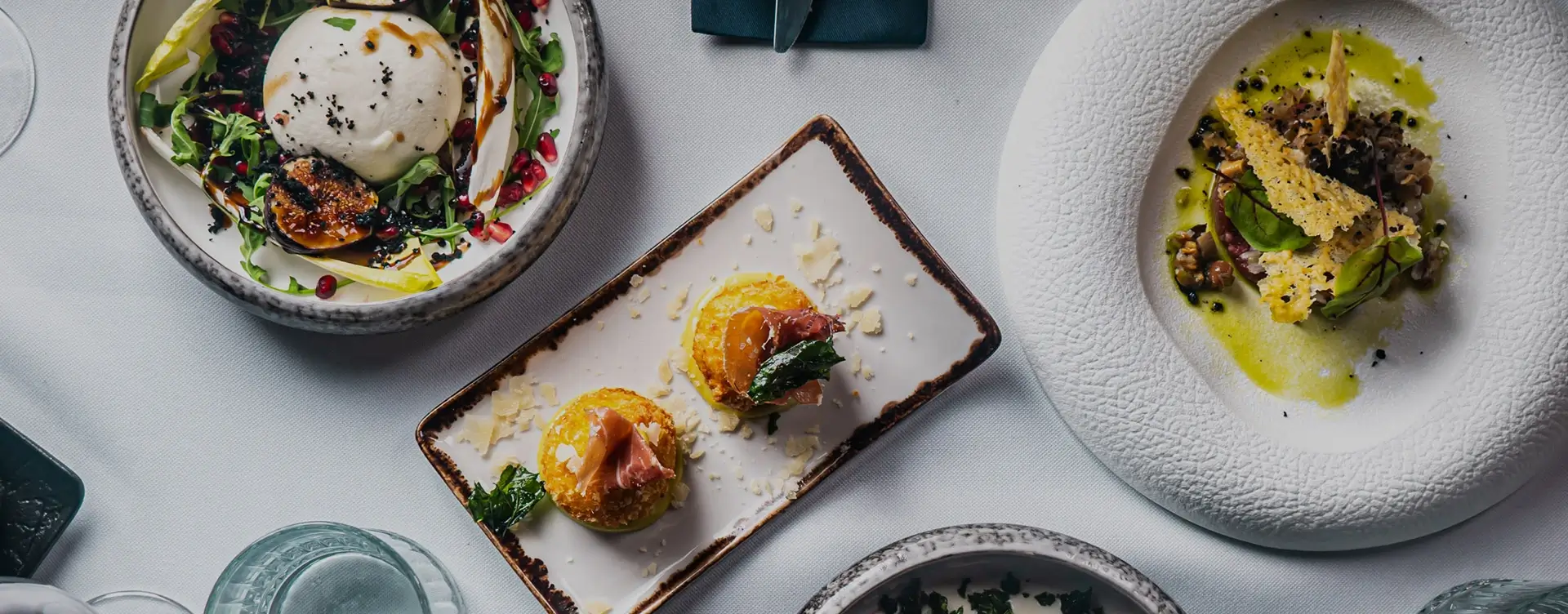 Elegancko nakryty stół z wykwintnymi przystawkami na białym obrusie, m.in. burratą z figami i owocami granatu oraz chrupiącymi roladkami z prosciutto, przygotowanymi na przyjęcie w restauracji Klub58.